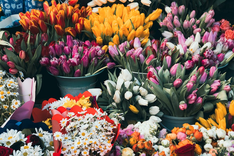 A vibrant collection of freshly cut tulips in various colors, including pink, white, orange, and yellow, arranged in metal buckets and wrapped bouquets. The tulips have smooth, elongated petals and st