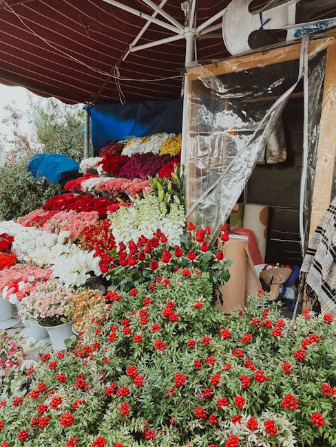A vibrant display of freshly cut flowers at a flower stall, arranged in neat layers with colors including white, pink, red, and deep burgundy. The flowers feature a mix of roses, chrysanthemums, and o