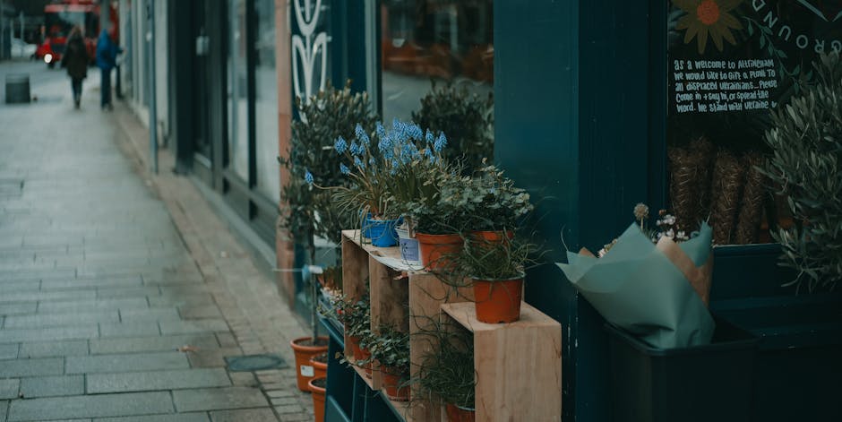 A close-up view of potted flowers arranged outdoors beside a storefront, with lavender and other small flowering plants in terracotta, blue, and beige pots placed on a wooden shelf. The arrangement is